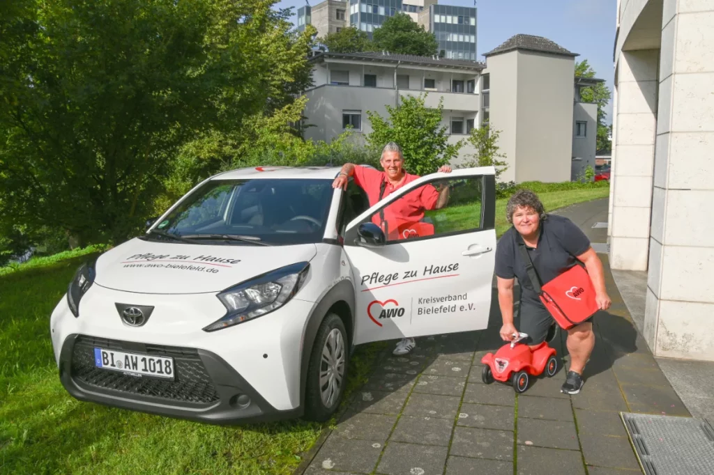 Zwei AWO-Mitarbeiterinnen stehen lächelnd vor einem weißen Dienstwagen der ambulanten Pflege – mit Tasche und rotem Bobbycar im Sonnenschein.